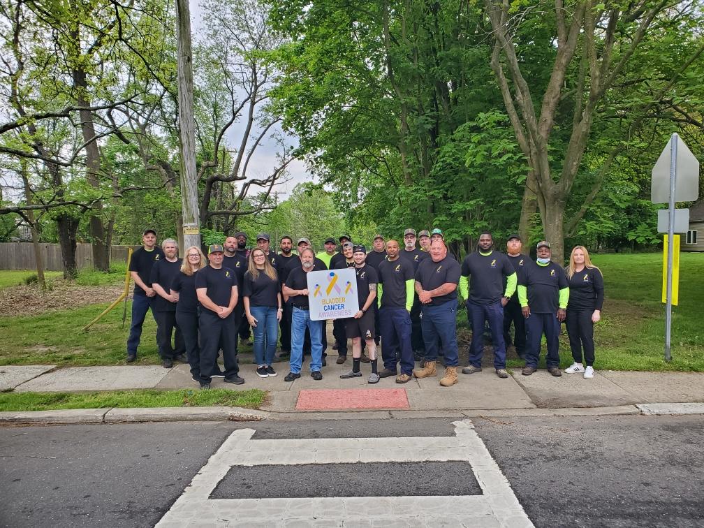 Group of volunteers in matching shirts poses outdoors in a park in front of a bladder cancer awareness sign.